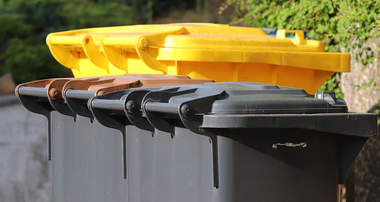 Eine Restabfalltonne mit gelben Deckel steht vor einem grünen Gebüsch